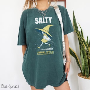 Pu&ograve; includere: T-shirt blu abete con la scritta "SALTY" sopra una grafica di una strega con cappello e vestito gialli. Il testo "LIBERAL WITCH RADICALIZED BY BASIC DECENCY" &egrave; sotto. La maglietta ha una vestibilit&agrave; ampia.