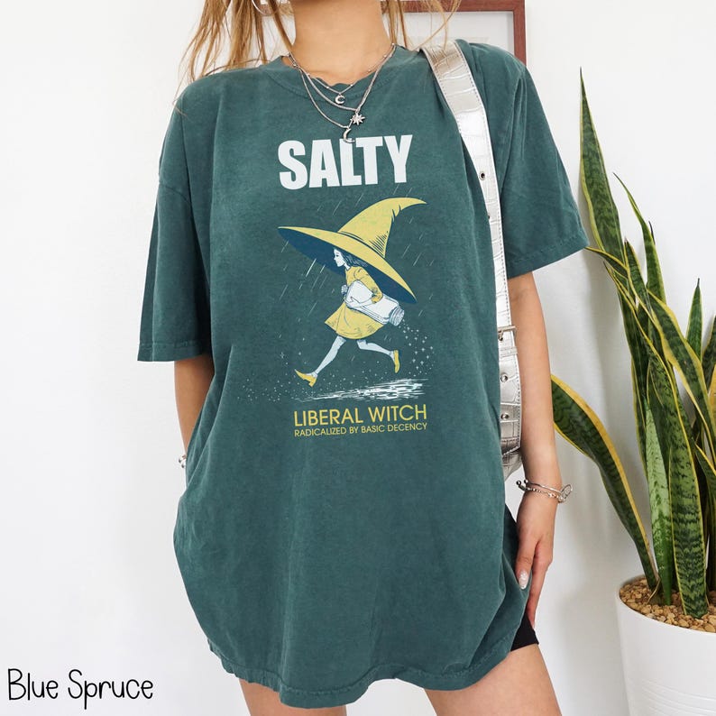Pu&ograve; includere: T-shirt blu abete con la scritta "SALTY" sopra una grafica di una strega con cappello e vestito gialli. Il testo "LIBERAL WITCH RADICALIZED BY BASIC DECENCY" &egrave; sotto. La maglietta ha una vestibilit&agrave; ampia.