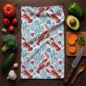 May include: A light blue kitchen towel with a repeating pattern of red lobsters, seashells, and blue coral. The towel is surrounded by fresh vegetables, including tomatoes, broccoli, and avocado, on a wooden surface.