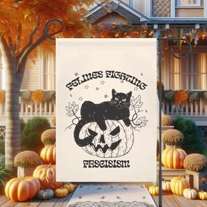 May include: A cream-colored garden flag with a black cat resting on a carved pumpkin. The flag reads "Felines Fighting Fascism" in a vintage font, with stars and leaves. The flag is displayed on a black metal stand, with pumpkins in the background.