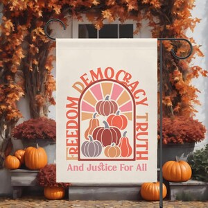 May include: A cream-colored garden flag with the words "Freedom, Democracy, Truth, And Justice For All" in a retro font. The flag features a pumpkin design in shades of orange, pink, and brown. The flag is displayed on a black metal stand, with pumpkins in the foreground.