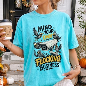 May include: A bright turquoise short-sleeve t-shirt featuring a graphic print of a security camera surrounded by five black crows. The text reads "MIND YOUR OWN FLOCKING BUSINESS" in bold black and yellow lettering. The shirt has a crew neckline.