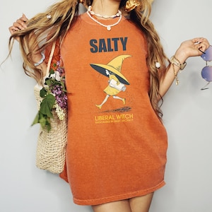 Pu&ograve; includere: Abito a maglietta arancione con la scritta "SALTY" in lettere maiuscole blu navy. La grafica presenta una strega che corre con un vestito e un cappello gialli, che tiene in mano una pergamena arrotolata. Il testo "LIBERAL WITCH" e "RADICALIZED BY BASIC DECENCY" sono sotto.
