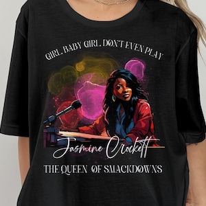 May include: Black t-shirt with a colorful graphic of a woman playing a keyboard. The text "Girl, Baby Girl, Don't Even Play" is above the graphic and "Jasmine Crockett The Queen of Smackdowns" is below the graphic.