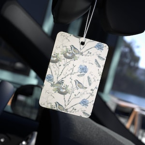 May include: A white car air freshener with a repeating pattern of blue birds, nests, and flowers. The text "First Time" is repeated throughout the design.