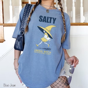 Pu&ograve; includere: Una maglietta blu jeans con la scritta "SALTY" sopra una grafica di una strega con cappello e vestito gialli. Il testo "LIBERAL WITCH RADICALIZED BY BASIC DECENCY" &egrave; sotto. La maglietta &egrave; uno stile casual e unisex.
