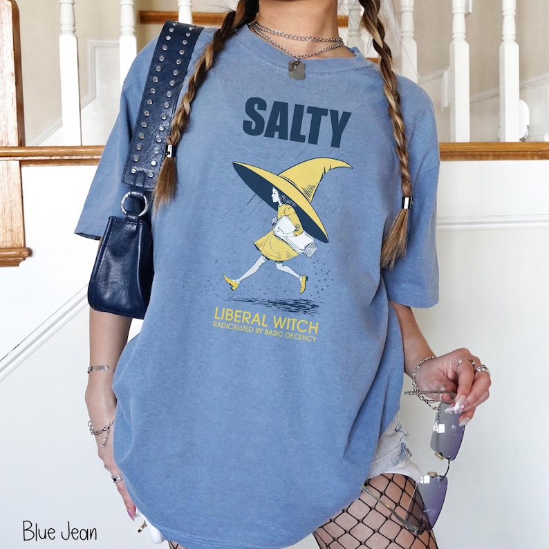 Pu&ograve; includere: Una maglietta blu jeans con la scritta "SALTY" sopra una grafica di una strega con cappello e vestito gialli. Il testo "LIBERAL WITCH RADICALIZED BY BASIC DECENCY" &egrave; sotto. La maglietta &egrave; uno stile casual e unisex.