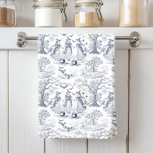 May include: White and blue patterned kitchen towel featuring a witch-themed toile design. The pattern includes witches, cauldrons, bats, trees, and spiderwebs. The towel is hanging on a silver bar.