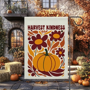 May include: A rectangular garden flag featuring the text HARVEST KINDNESS in a bold brown font. The design displays a large orange pumpkin centered at the bottom, surrounded by stylized flowers and leaves in shades of orange, yellow, and brown.