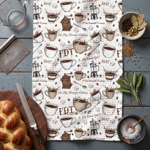 May include: A white waffle-weave kitchen towel featuring a repeating pattern of coffee cups, French presses, coffee grinders, and sugar cubes. The text includes FDT, 8647, Is He Dead Yet?, and The Big Beautiful Obituary, F*ck Donald Trump.