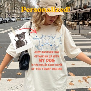 May include: Cream t-shirt featuring a blue dog illustration and the text "JUST ANOTHER DAY OF WAKING UP WITH MY DOG in the upside down world OF THE TRUMP REGIME." A photo of a dog is attached. The word "Personalized!" is at the top.