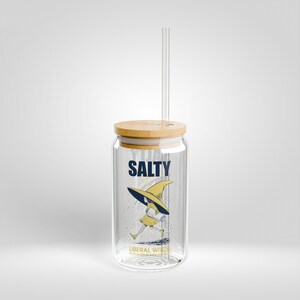 May include: A clear glass can-shaped drinking glass with a wooden lid and a clear straw. The glass features the word "SALTY" in navy blue, and a cartoon of a running witch in yellow and navy blue. The text "LIBERAL WITCH" is below the cartoon.