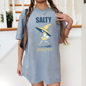 Pu&ograve; includere: T-shirt azzurra con la scritta "SALTY" sopra una grafica di una strega che corre sotto la pioggia. La strega indossa un vestito e un cappello gialli. Il testo "LIBERAL WITCH RADICALIZED BY BASIC DECENCY" &egrave; sotto.
