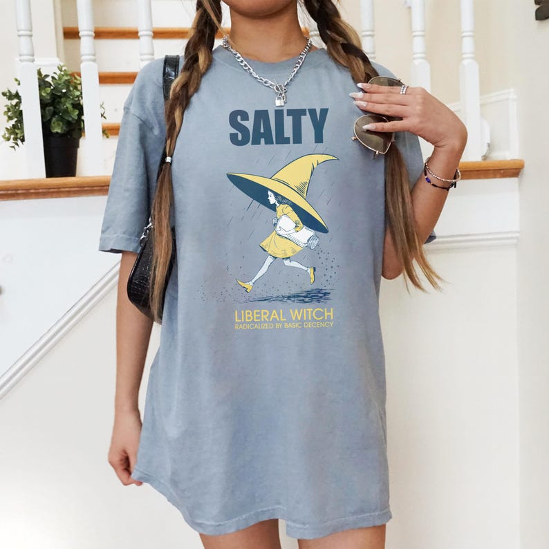 Pu&ograve; includere: T-shirt azzurra con la scritta "SALTY" sopra una grafica di una strega che corre sotto la pioggia. La strega indossa un vestito e un cappello gialli. Il testo "LIBERAL WITCH RADICALIZED BY BASIC DECENCY" &egrave; sotto.