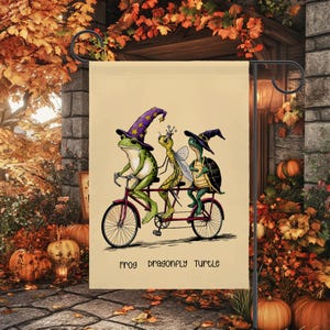 May include: A garden flag featuring a whimsical illustration of a frog, dragonfly, and turtle riding a tandem bicycle. The frog wears a wizard hat, the dragonfly a crown, and the turtle a witch hat. The text "FROG DRAGONFLY TURTLE" is at the bottom.