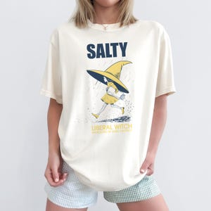 Pu&ograve; includere: T-shirt color crema con la scritta "SALTY" in blu navy a blocchi. Sotto, una strega dei cartoni animati con un vestito e un cappello gialli corre sotto la pioggia. Il testo "LIBERAL WITCH" &egrave; sotto l'immagine, con "RADICALIZED BY BASIC DECENCY" sotto.