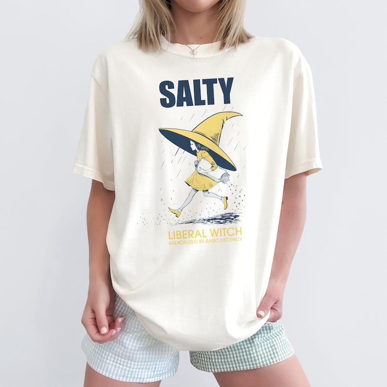Pu&ograve; includere: T-shirt color crema con la scritta "SALTY" in blu navy a blocchi. Sotto, una strega dei cartoni animati con un vestito e un cappello gialli corre sotto la pioggia. Il testo "LIBERAL WITCH" &egrave; sotto l'immagine, con "RADICALIZED BY BASIC DECENCY" sotto.
