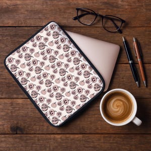 May include: A laptop sleeve with a black border and a repeating pattern of smiling pumpkins in shades of pink and cream. The sleeve is on a wooden desk with a laptop, glasses, pens, and a cup of coffee.