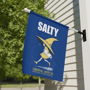 May include: A blue flag with the word "SALTY" in white at the top. Below is an illustration of a witch in a yellow dress and hat, carrying a salt shaker. The text "LIBERAL WITCH RADICALIZED BY BASIC DECENCY" is at the bottom.