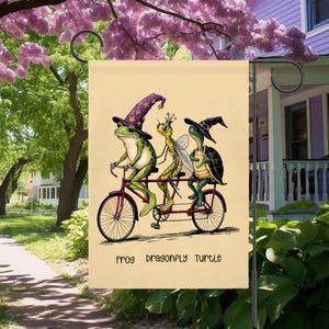 May include: A whimsical garden flag features a frog, dragonfly, and turtle riding a tandem bicycle. The frog wears a wizard hat, the dragonfly a crown, and the turtle a witch hat. The flag has the words "FROG DRAGONFLY TURTLE" at the bottom.