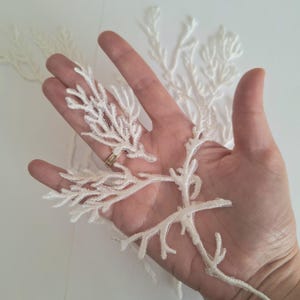 May include: Ivory lace appliques with a botanical design, similar to coral or branches. The appliques are held in a hand, displaying the detailed embroidery and texture. Perfect for embellishing clothing or craft projects.