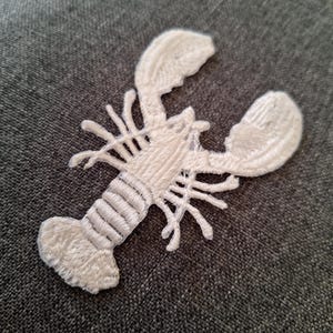 May include: A white embroidered lobster patch. The patch features a detailed design of a lobster with claws, legs, and a segmented body. The lobster is made of white thread and is set against a gray fabric background.