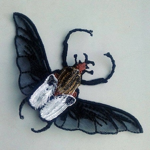 Könnte beinhalten: Gestickte Insektenapplikation mit schwarzen, silbernen, goldenen und roten Akzenten. Das Insekt hat große, detaillierte Flügel und einen strukturierten Körper. Diese dekorative Applikation kann für Kleidung, Accessoires oder Bastelprojekte verwendet werden.
