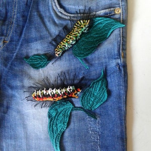 May include: Blue denim jeans with two embroidered patches of caterpillars on green leaves. The caterpillar on the top is green and yellow, and the caterpillar on the bottom is black, red, white, and yellow.