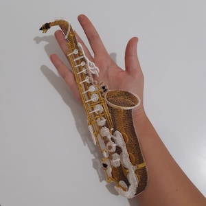 以下が含まれることがあります： 金、茶色、白の刺繍サックスパッチ。パッチは、楽器のキーとベルを示す詳細なステッチが特徴です。パッチは、生地にアイロンをかけるか縫い付けるように設計されています。