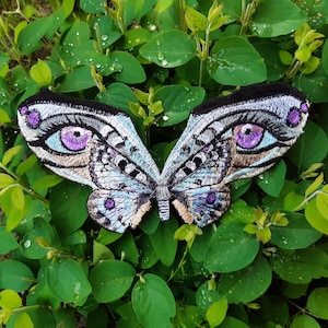 May include: A blue and silver embroidered butterfly patch with purple eyes. The patch is on a bed of green leaves.