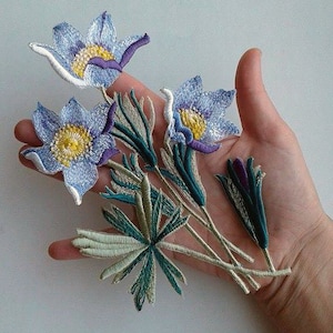 May include: A set of five embroidered flower patches featuring blue and purple flowers with yellow centres and green leaves.