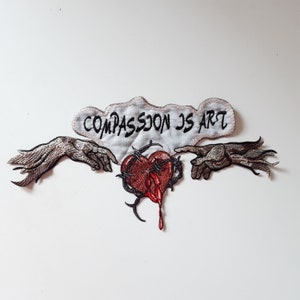 Peut inclure: Patch brodé avec deux mains qui tendent vers un cœur saignant avec des épines. Le texte "COMPASSION IS ART" est sur un nuage blanc au-dessus des mains.