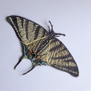 Op de afbeelding: Een geborduurde vlinderpatch in zwart en goud met turquoise accenten. De vlinder bevindt zich in een vliegende positie met uitgespreide vleugels.