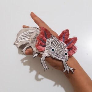 May include: Embroidered axolotl patch with pink and purple frills, white and gray body, and blue eyes.