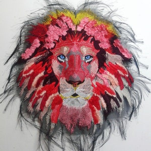 Puede incluir: Cabeza de león bordada con una melena roja, rosa y amarilla. Los ojos del león son azules y el fondo es negro.