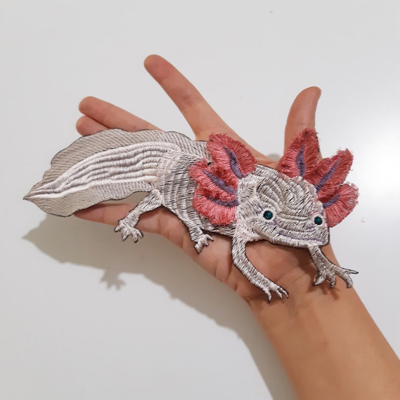 May include: A white axolotl with pink feathery gills, embroidered on a fabric patch. The axolotl is facing the viewer with its mouth slightly open.