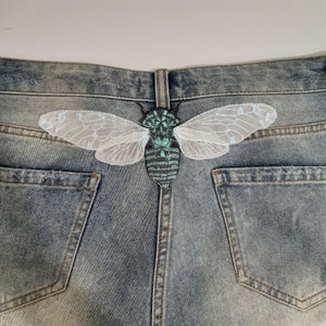 Pu&ograve; includere: Toppa ricamata a forma di cicala con dettagli turchese e grigio, e delicate ali in pizzo bianco, applicata sul retro di un paio di pantaloncini di jeans blu. La toppa &egrave; centrata sulla cintura, aggiungendo un elemento decorativo unico.