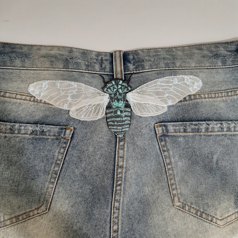 Pu&ograve; includere: Toppa ricamata a forma di cicala con dettagli turchese e grigio, e delicate ali in pizzo bianco, applicata sul retro di un paio di pantaloncini di jeans blu. La toppa &egrave; centrata sulla cintura, aggiungendo un elemento decorativo unico.