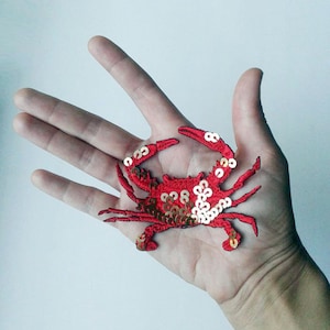 May include: A red crab-shaped embroidered patch with gold sequins. The crab has detailed stitching and is displayed on a light background. This decorative patch is suitable for clothing and accessories.