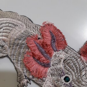 May include: Embroidered iron-on patch of an axolotl, a type of salamander, with pink feathery gills and a green eye. The axolotl is white with black and silver embroidery.
