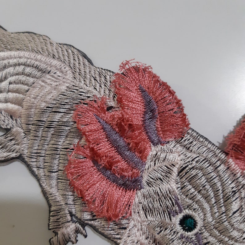 May include: Embroidered iron-on patch of an axolotl, a type of salamander, with pink feathery gills and a green eye. The axolotl is white with black and silver embroidery.