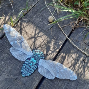 Puede incluir: Una cigarra bordada con un cuerpo iridiscente en azul turquesa y azul marino y delicadas alas blancas transparentes. El insecto está sobre tablas de madera desgastadas, con hierba en el fondo.