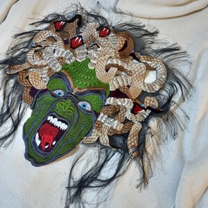 May include: A white sweater with a green and brown embroidered patch of Medusa's head. The patch features a snarling face with red lips and blue eyes, surrounded by snakes with red eyes.