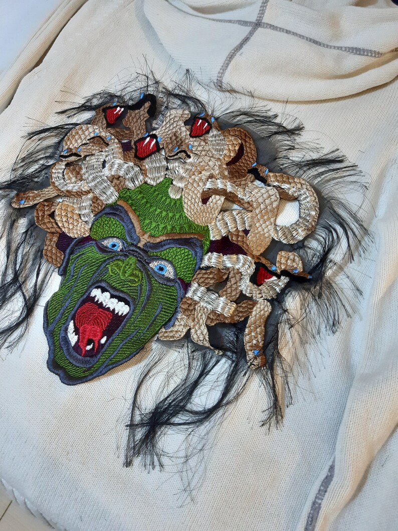 May include: A white sweater with a green and brown embroidered patch of Medusa's head. The patch features a snarling face with red lips and blue eyes, surrounded by snakes with red eyes.