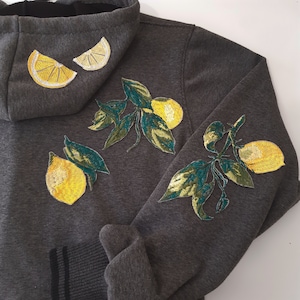 Può includere: Una felpa con cappuccio grigio scuro con disegni di limone ricamati sul davanti e sulle maniche. I limoni sono gialli con foglie verdi.