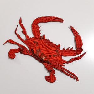 May include: Red embroidered crab applique with gold sequins. This iron-on patch is perfect for adding a nautical touch to clothing, bags, or home decor.
