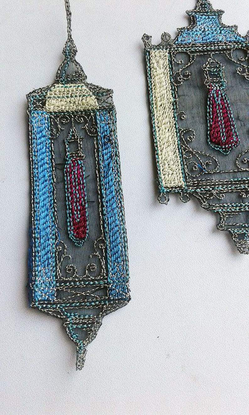 K&ouml;nnte beinhalten: Zwei verzierte, bestickte Stoffst&uuml;cke. Das Hauptst&uuml;ck hat eine rechteckige Form mit blauem Rand und einem zentralen Design in Rot und Blau. Das zweite St&uuml;ck ist &auml;hnlich, mit einem cremefarbenen Rand und einem rot-blauen Design.