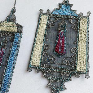 K&ouml;nnte beinhalten: Zwei verzierte, rechteckige Dekorationen mit aufwendigen silberfarbenen Drahtdetails. Jedes hat ein zentrales Design mit einer roten und blauen Quaste, umrahmt von cremefarbener und blauer Stickerei. Die St&uuml;cke haben eine Vintage- und handwerkliche &Auml;sthetik.