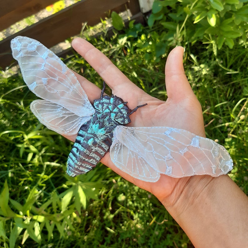 Pu&ograve; includere: Una decorazione artigianale a forma di insetto, una cicala, con corpo iridescente turchese e nero, e delicate ali di pizzo bianco. L'insetto &egrave; tenuto in mano su uno sfondo di erba verde e luce solare.