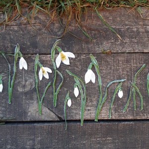 May include: A collection of white and green felt snowdrop flower appliques. The appliques are arranged in a row on a wooden surface.